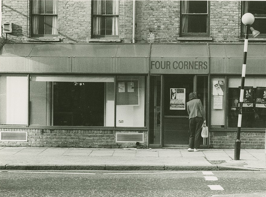 A black and white archive photograph of Four Corners building, taken around 1970. A man is entering the building. There is a poster on the glass door. 