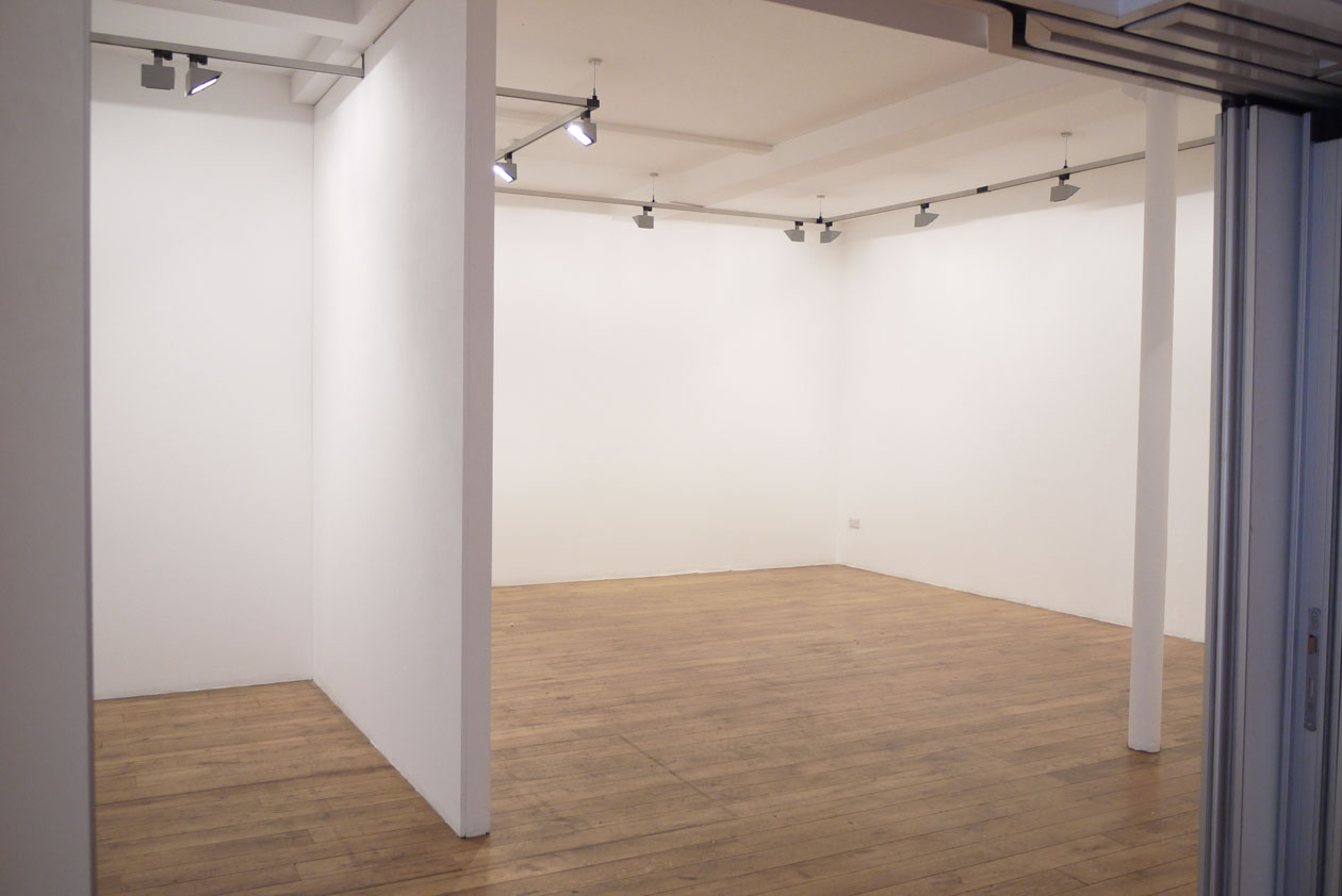 Four Corners' empty gallery with white walls and wooden floors, split into two sections, with a pillar on the right hand side.