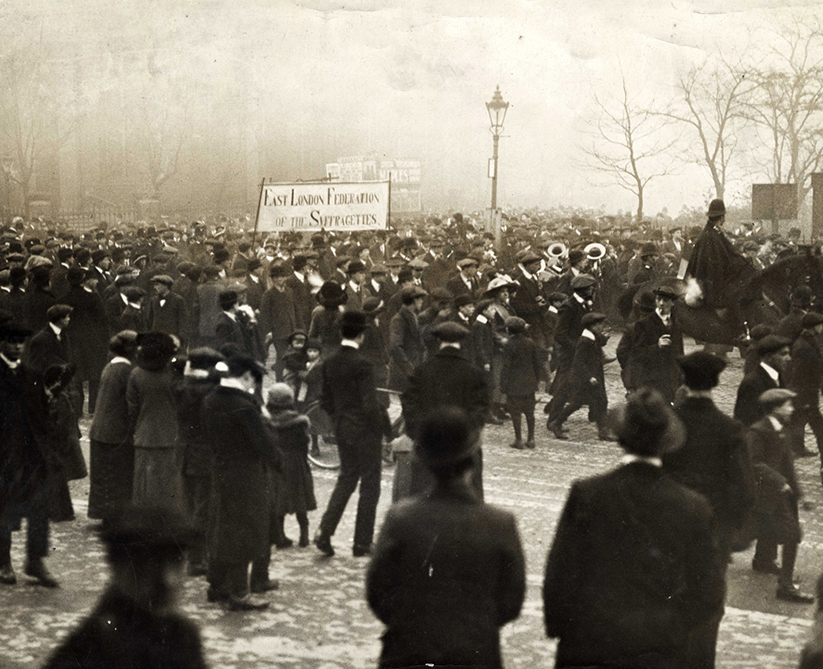 Black and white archive photograph of an East End Federation of Suffragettes march in Victoria Park. It it misty and the trees are bare. There is a mounted police officer to the right. 