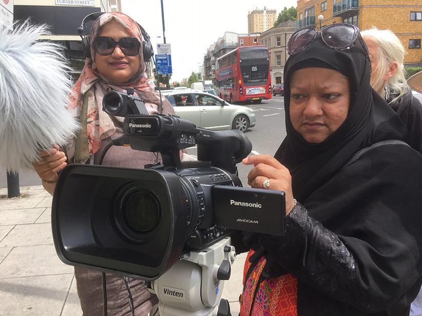 Two women in headscarves stand in the street holding a film camera and boom mic. 