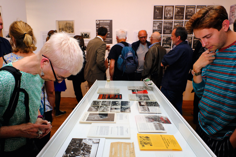 A busy gallery. In the foreground are two people looking into a display case with paper documents inside.