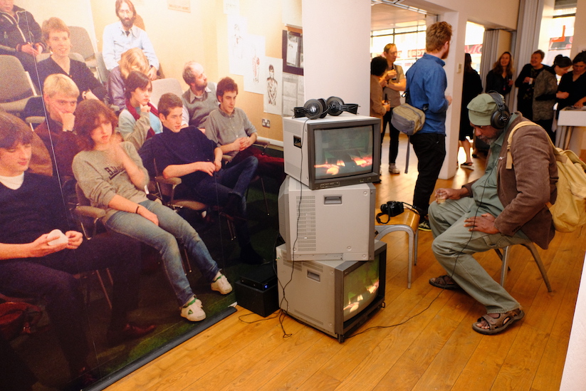 A man sitting on a chair in a gallery, watching a film on an old fashioned television set. 