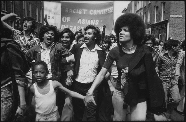 Black and white photograph shows a lively protest. In the foreground a woman with an afro holds the hand of a small boy. The protesters behind her are chanting. A banner in the background reads: Anti-Racist committee of Asians in East London.