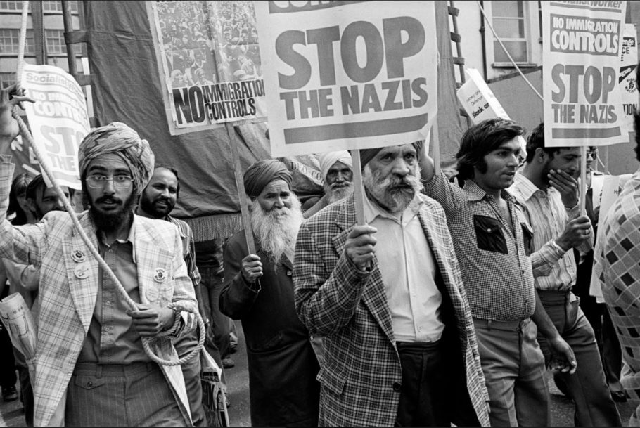 Black and white photograph of men protesting. Many are wearing turbans and have beards. They are holding placards that read, No immigration controls. Stop the Nazis