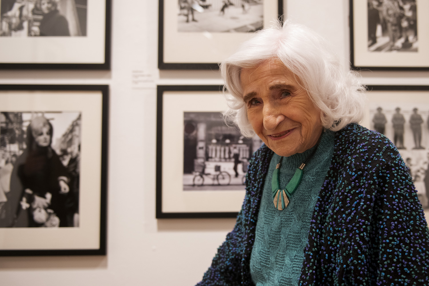 The photographer Dorothy Bohm stands in front of her framed photographs, smiling
