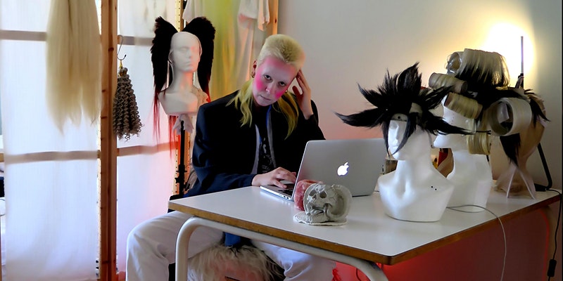  A person with bright yellow hair and striking pink and white face makeup sits at a desk behind an Apple Mac computer. They are touching the side of their head with one hand. They are flanked by two white busts  dressed in elaborate feather headdresses.