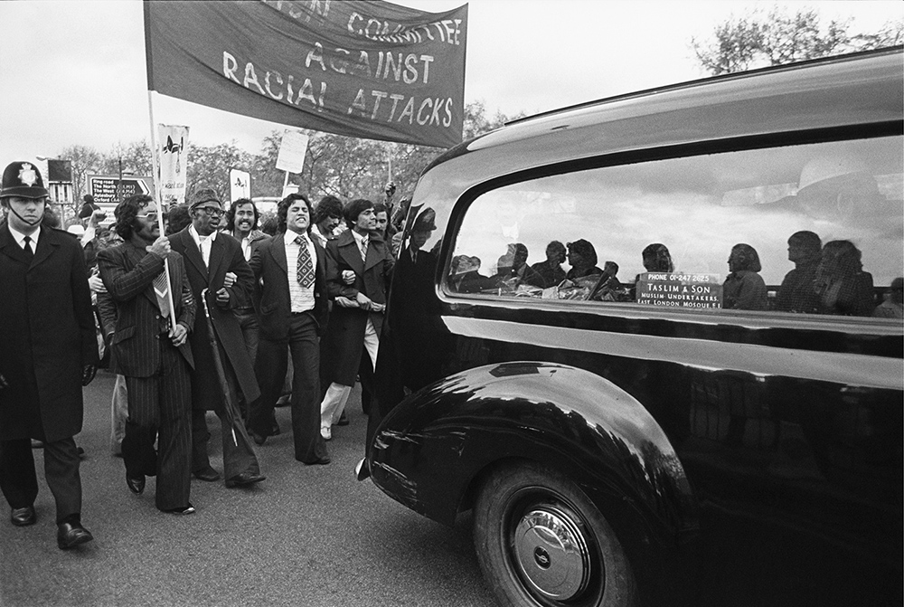 Black and white photograph of people marching behind Altab Ali's coffin to Downing Street,