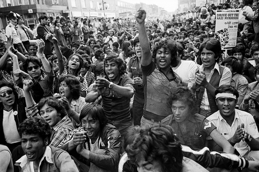 Black and white photograph of large crowd at Brick Lane 1978 protests. A man in the centre holds up his fist.