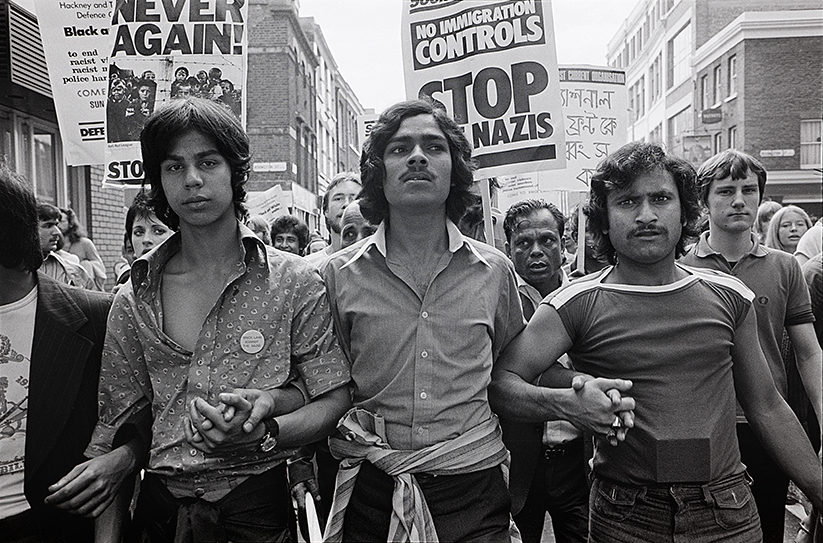 Black and white photograph. Three Bengali men with linked arms lead a crowd of protesters. Placards in the background read No Immigration Controls, Stop the Nazis