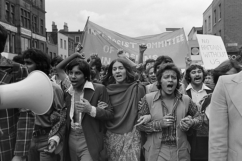 Two men and one woman with arms linked lead a crowd of protesters. They are shouting. Banners and placards are raised in the distance.