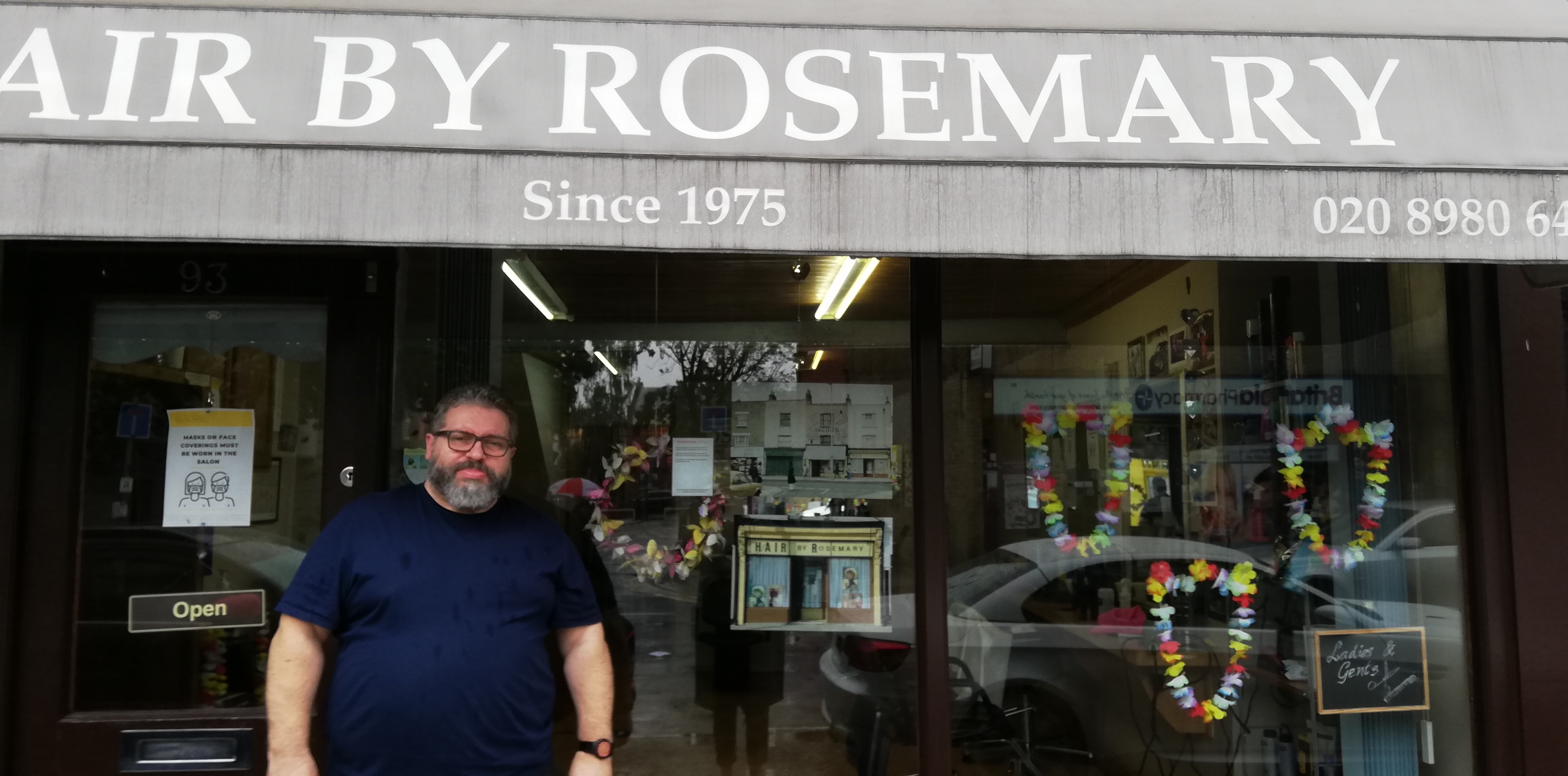 A man in a blue shirt is smiling outside a hairdresser. There is an old photograph of the same hairdresser displayed in the window