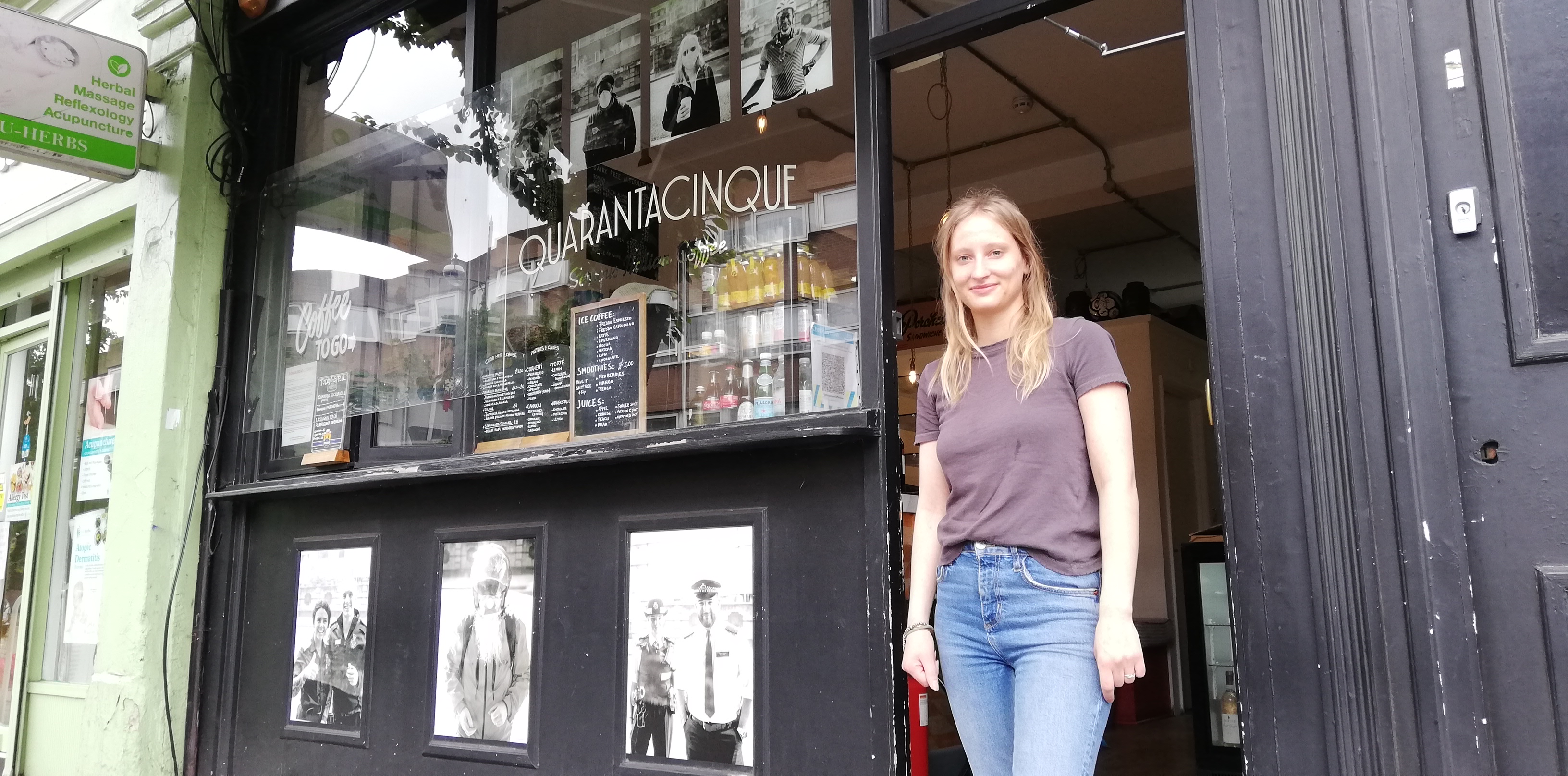 A woman smiles in the doorway of a coffee shop. There are large black and white prints displayed on the front of the building. 