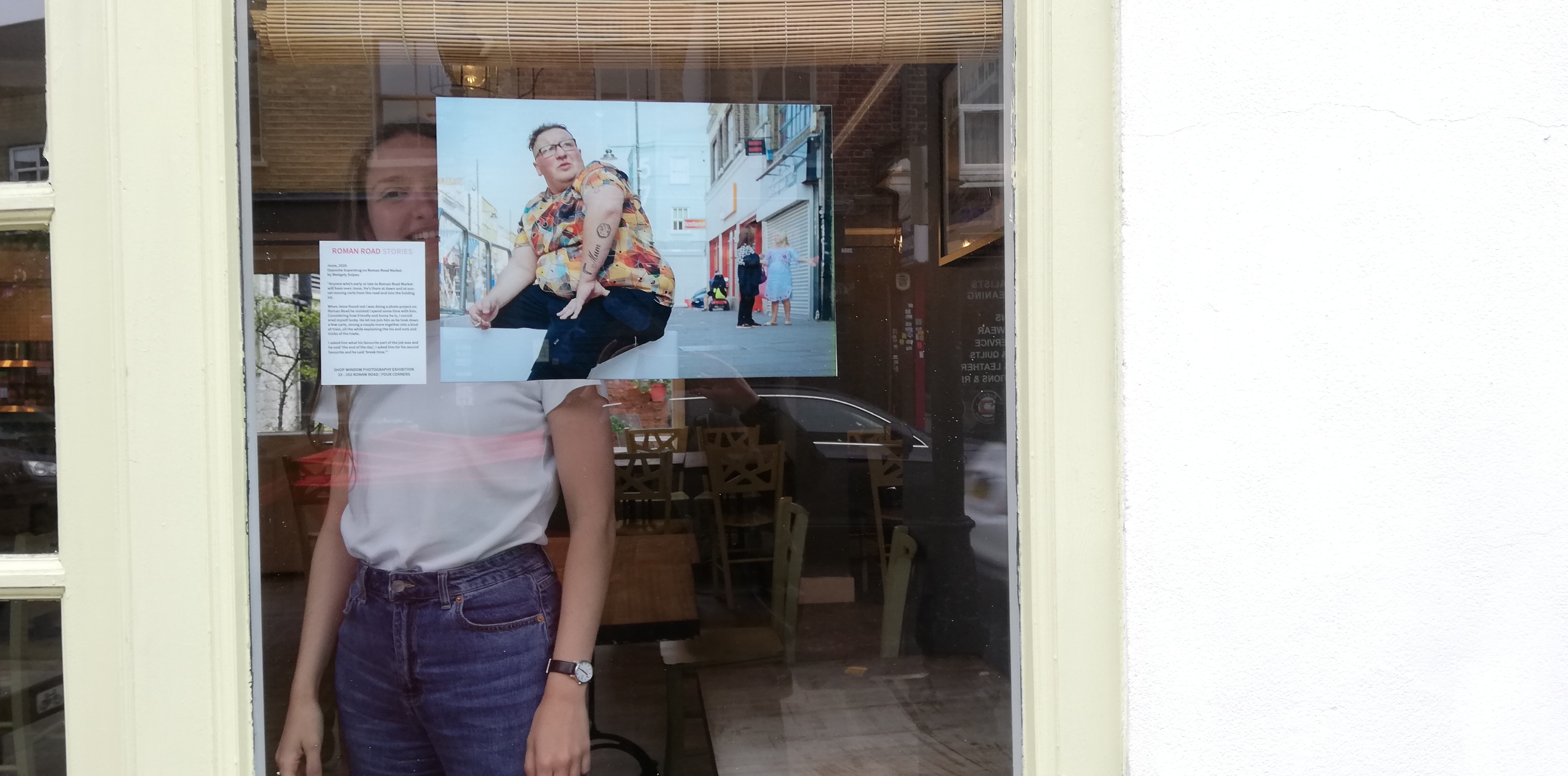 A woman behind the glass window of a cafe. In the window is a photograph of a man in a colourful shirt 