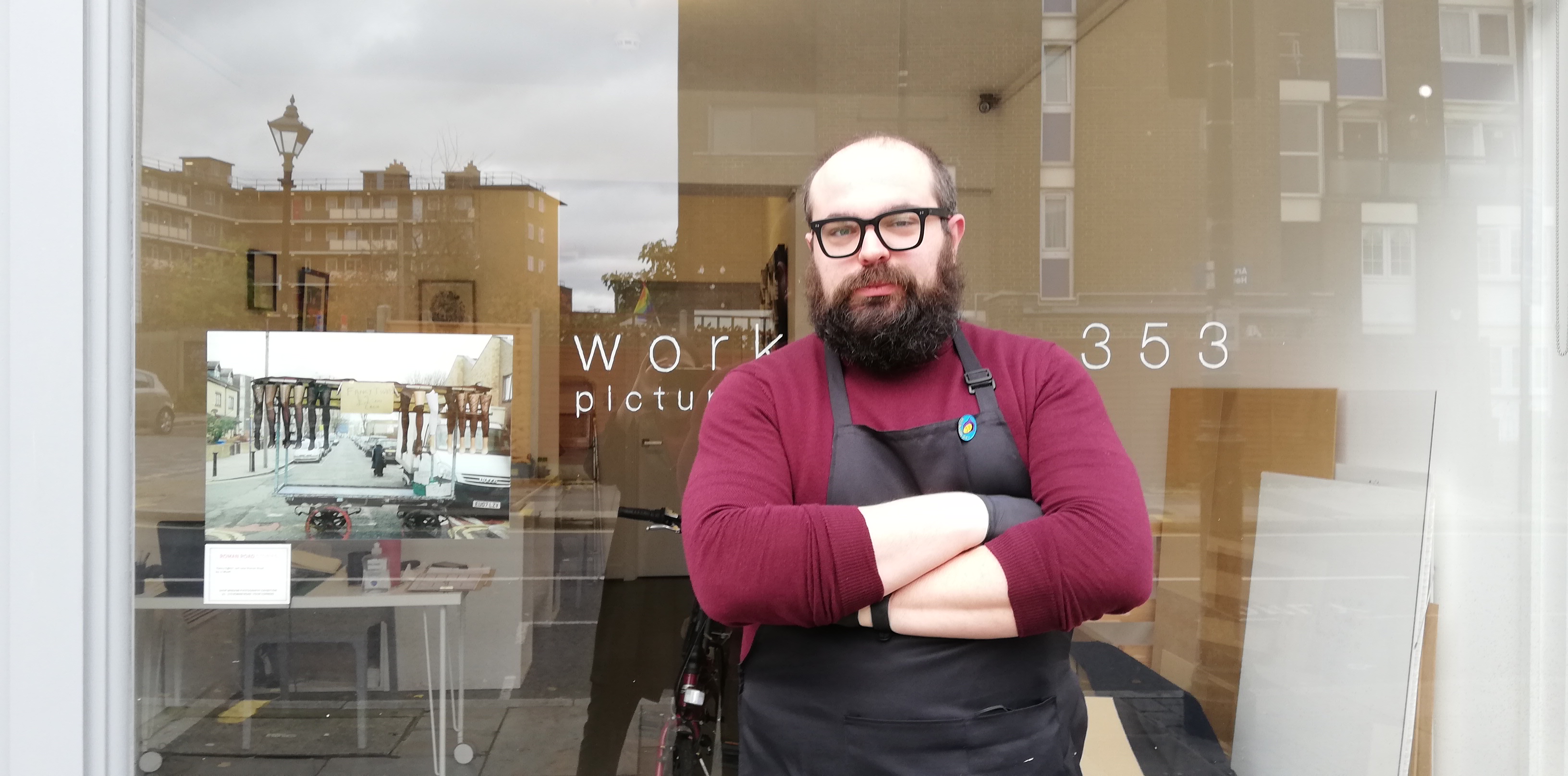 A framer stands outside his shop. There is a photograph of a market stall in the window. 