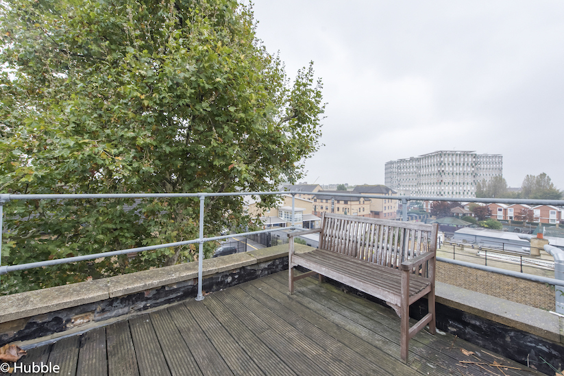 Roof terrace with views of London. The terrace has dark deck flooring and a wooden bench, with metal railings. There is a tree nearby.